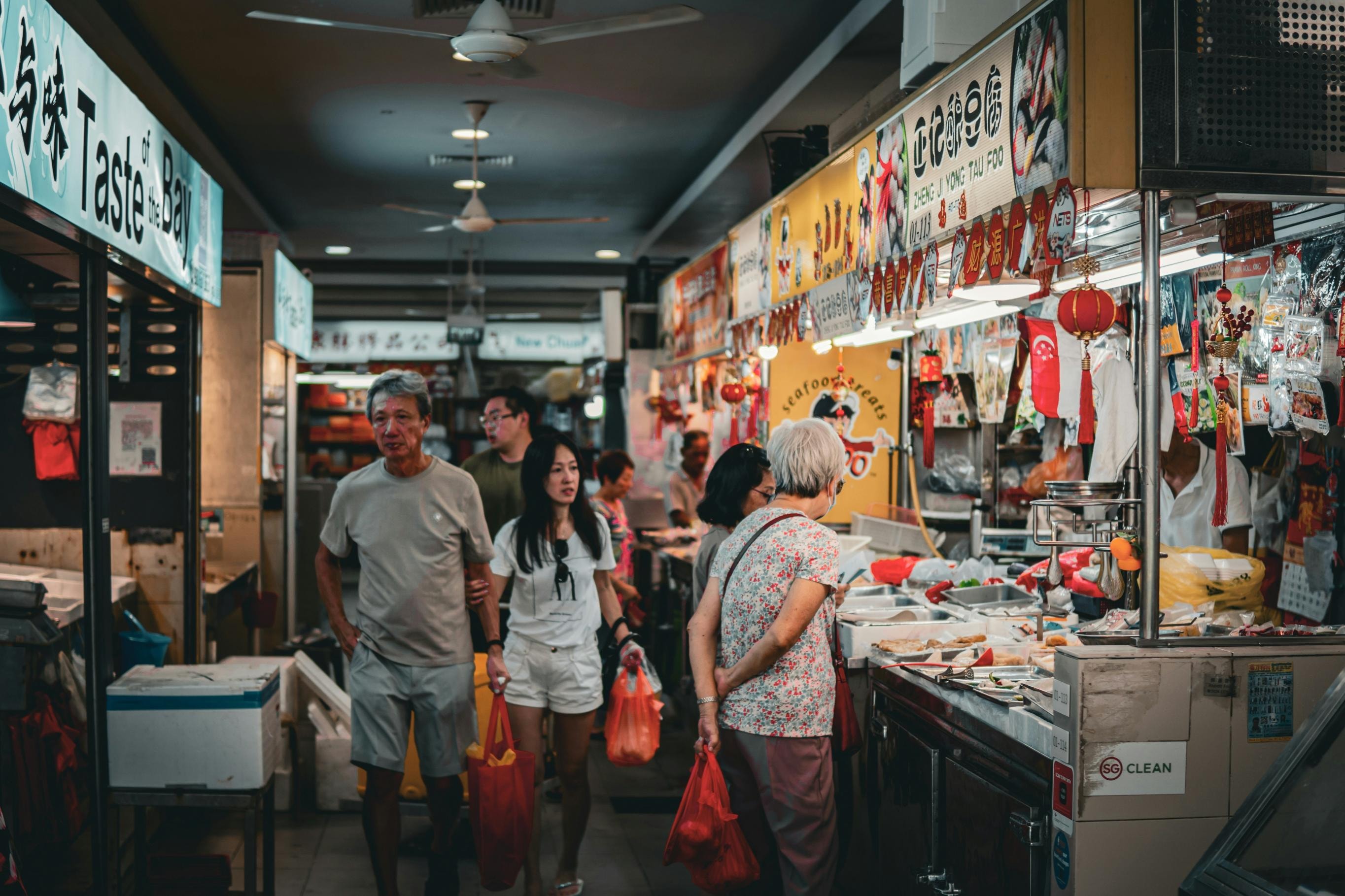 This image captures the busy interior of a Singaporean hawker center, where patrons carry bags of food past various stalls. The scene is filled with a variety of colorful signage, hanging decorations, and the lively energy of a local food market.