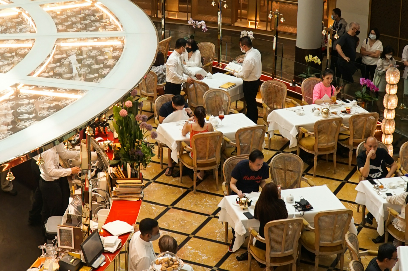 An elegant restaurant scene with diners seated at white tablecloth tables. Waitstaff in white uniforms serve guests. Soft lighting and floral decor create a cozy ambiance.