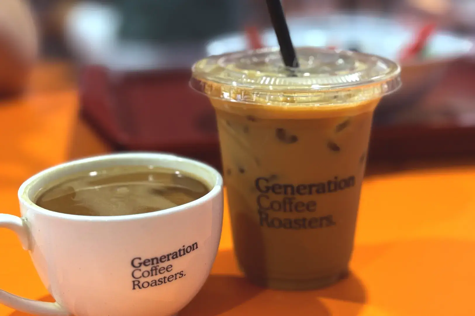A hot ceramic cup of coffee next to a cold iced coffee in a plastic takeaway cup, both branded with the Generation Coffee Roasters logo.