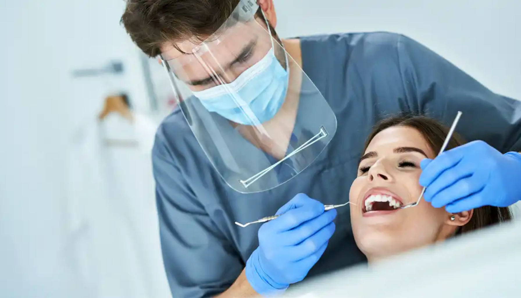 Tannlege med visir og munnbind undersøker kvinnelig pasient med speil og sonde under rutinekontroll i en moderne tannklinikk.