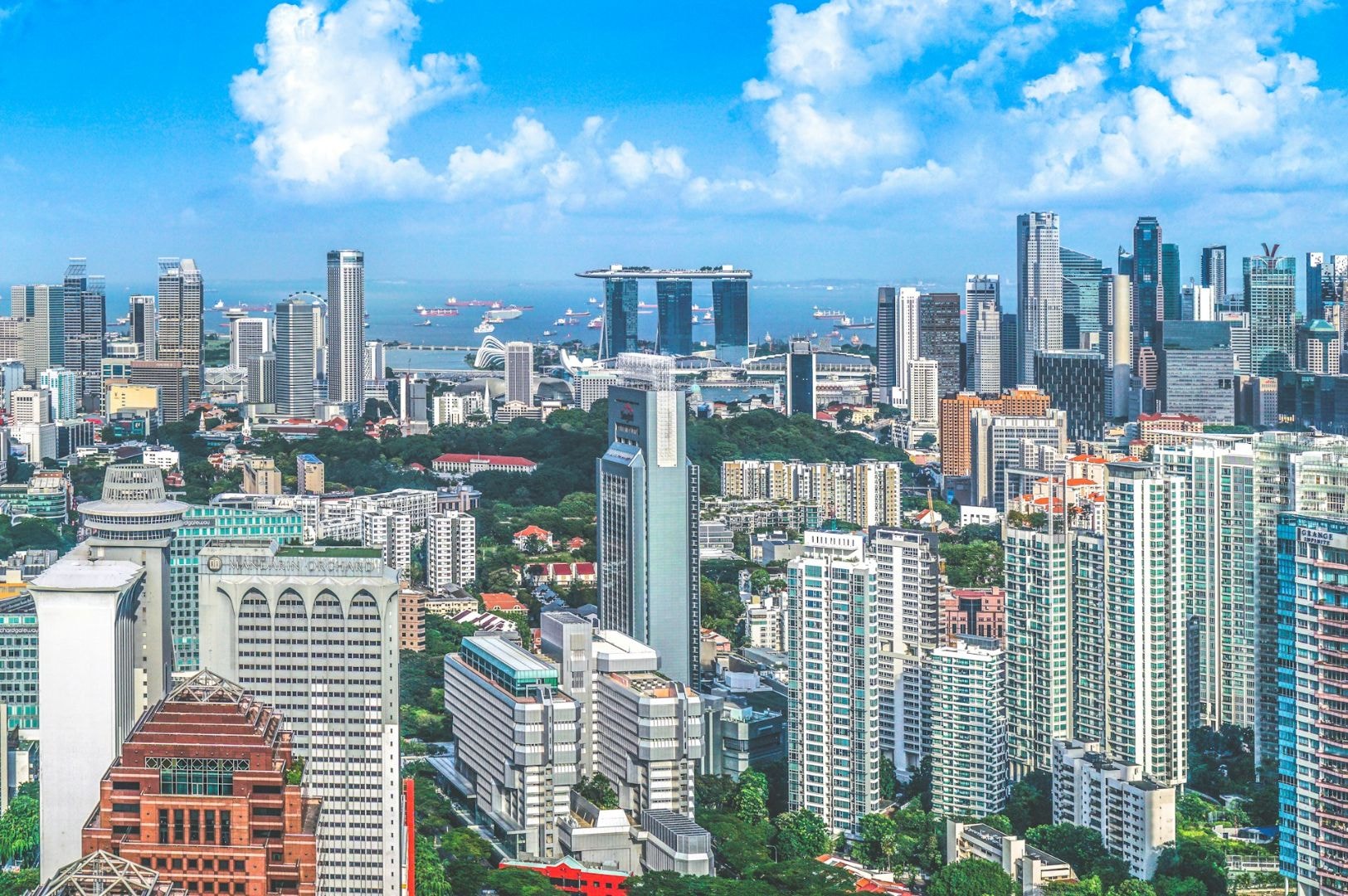 Aerial view of Singapore's skyline with modern skyscrapers, including a unique boat-shaped building. Ocean and ships visible under a bright blue sky.