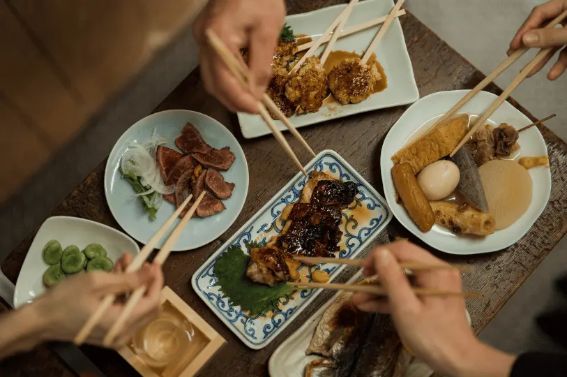 Top-down view of diners' hands using chopsticks to share a feast of Japanese dishes, including yakitori, oden, and sliced meats, at a rustic wooden table in a Singapore izakaya.