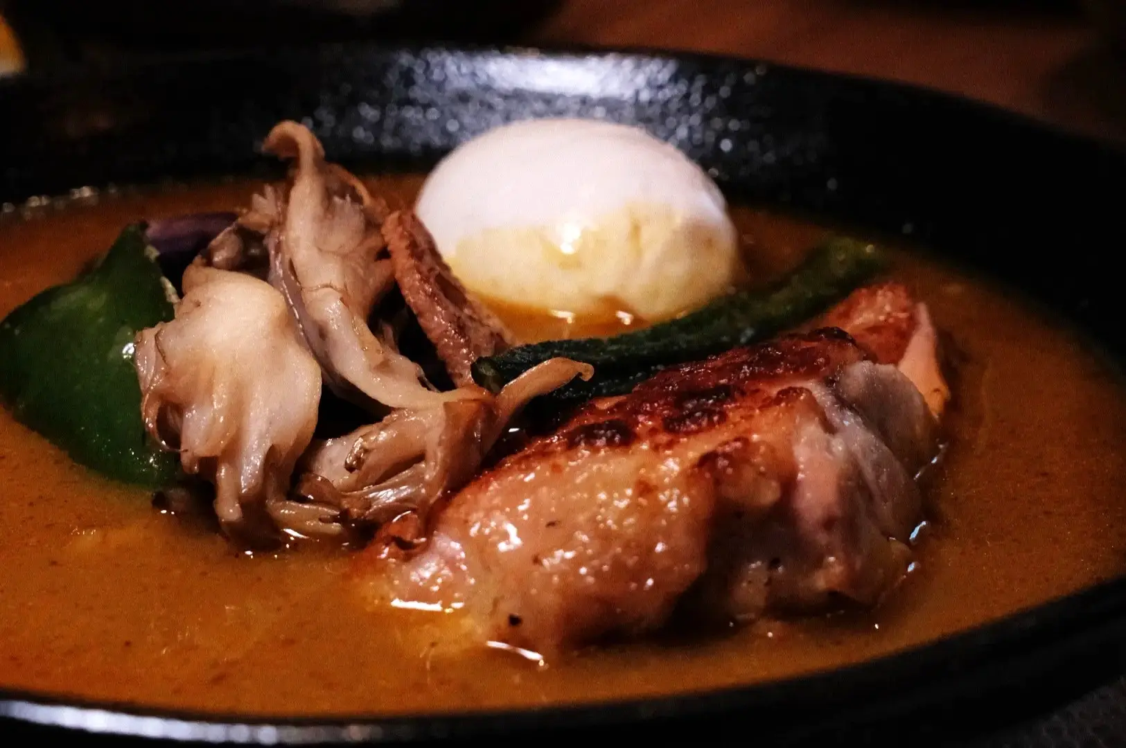 A close-up of a dish featuring grilled chicken, assorted mushrooms, green peppers, and a poached egg in a savory broth.