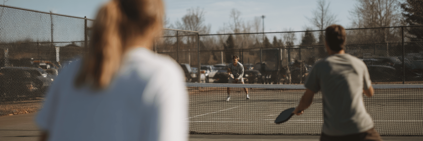 Two adults playing pickleball on a sunlit outdoor court, one swinging a paddle as the ball crosses the net.