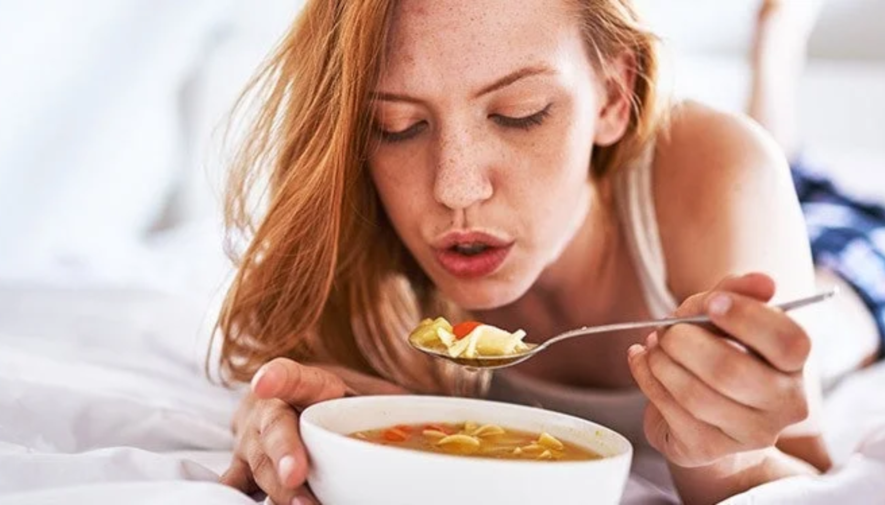 Rødhåret kvinne ligger i sengen og blåser på en skje med varm suppe.
