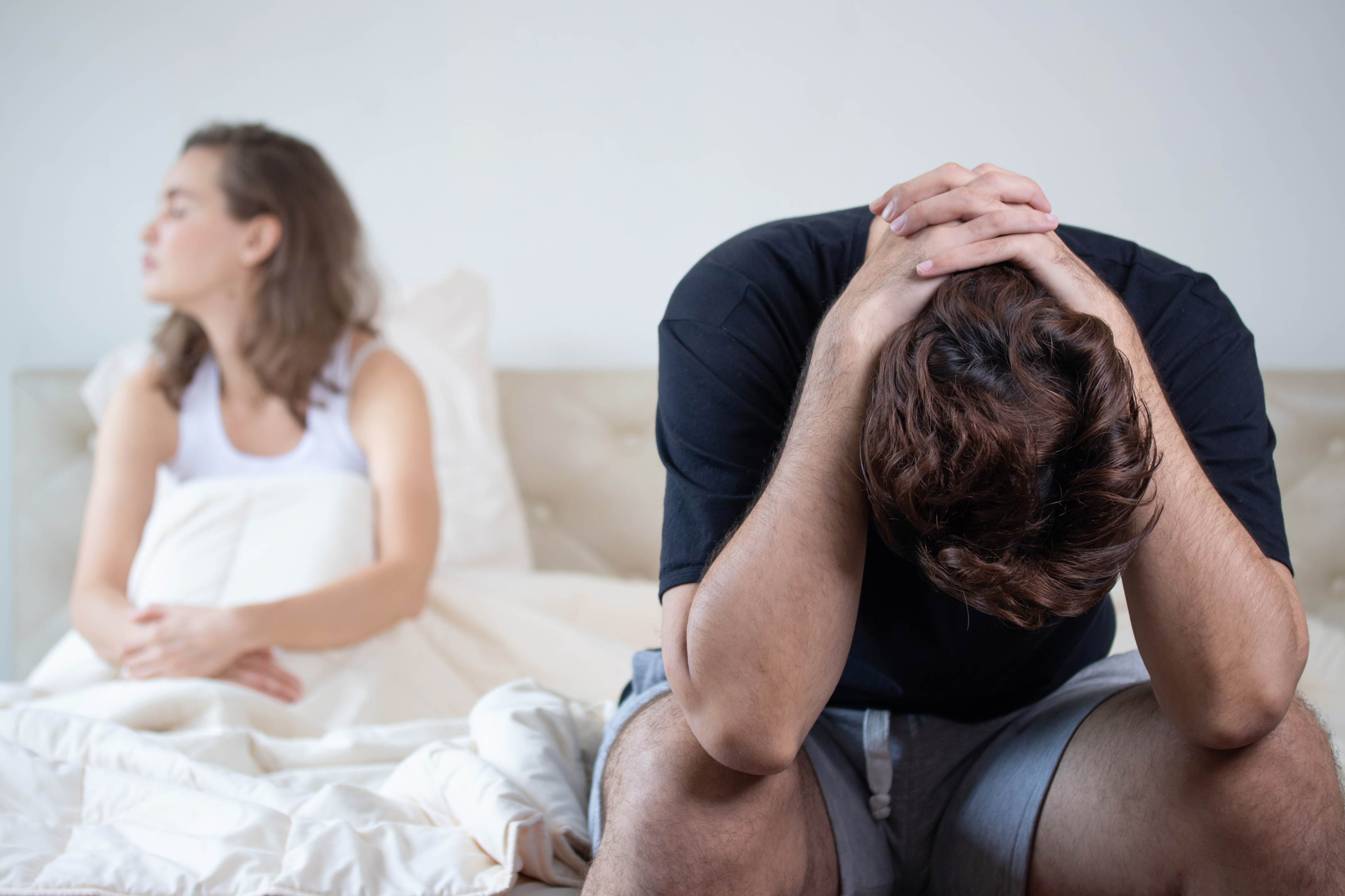 Image shows a caucasian couple, her on the left in the distance, long brown wavy hair, covers pulled up over her knees. The male is in the foreground, sitting on the edge of the bed with his elbows on his knees and his hands clasped behind his head which is facing the floor. He has dark hair and is wearing a black t-shirt.