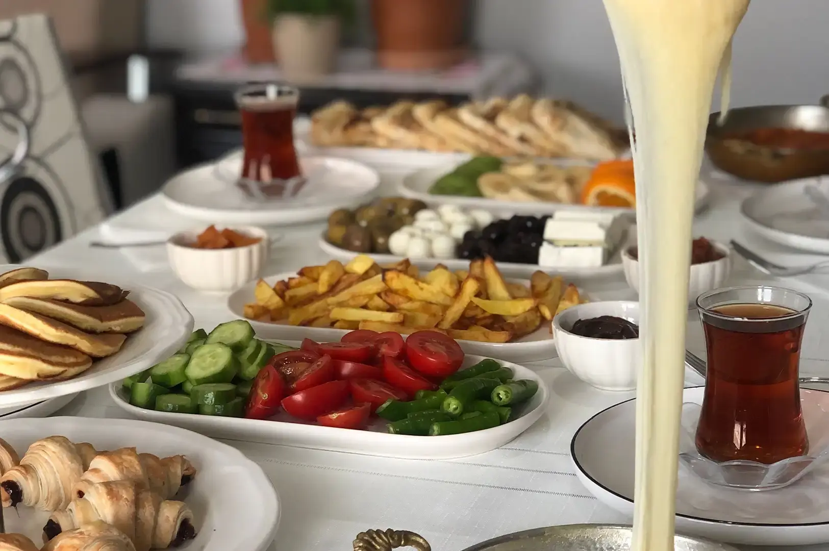 A lavish Turkish breakfast spread covers a white table, featuring sliced cucumbers, tomatoes, peppers, french fries, pastries, pancakes, olives, cheeses, and glasses of Turkish tea, with a tall, dramatic stream of melted cheese being stretched upwards in the foreground.