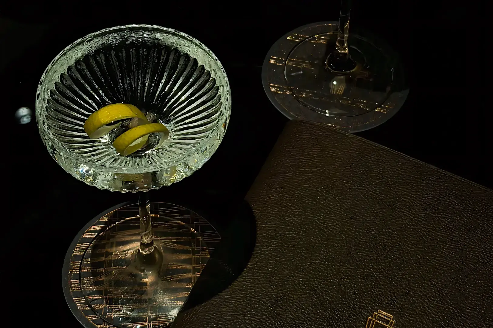 Overhead top‑down shot of a crystal cocktail glass garnished with lemon peels beside a leather‑bound menu on a dark tabletop, highlighting Atlas Bar Singapore’s refined cocktail presentation.
