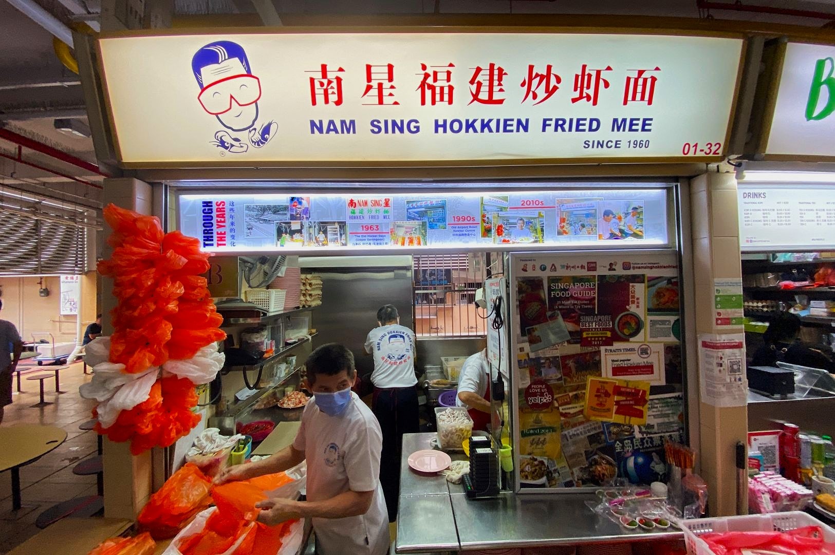Hawker stall named "Nam Sing Hokkien Fried Mee" with signage in English and Chinese. Cook in white shirt and mask prepares food. Casual and bustling.