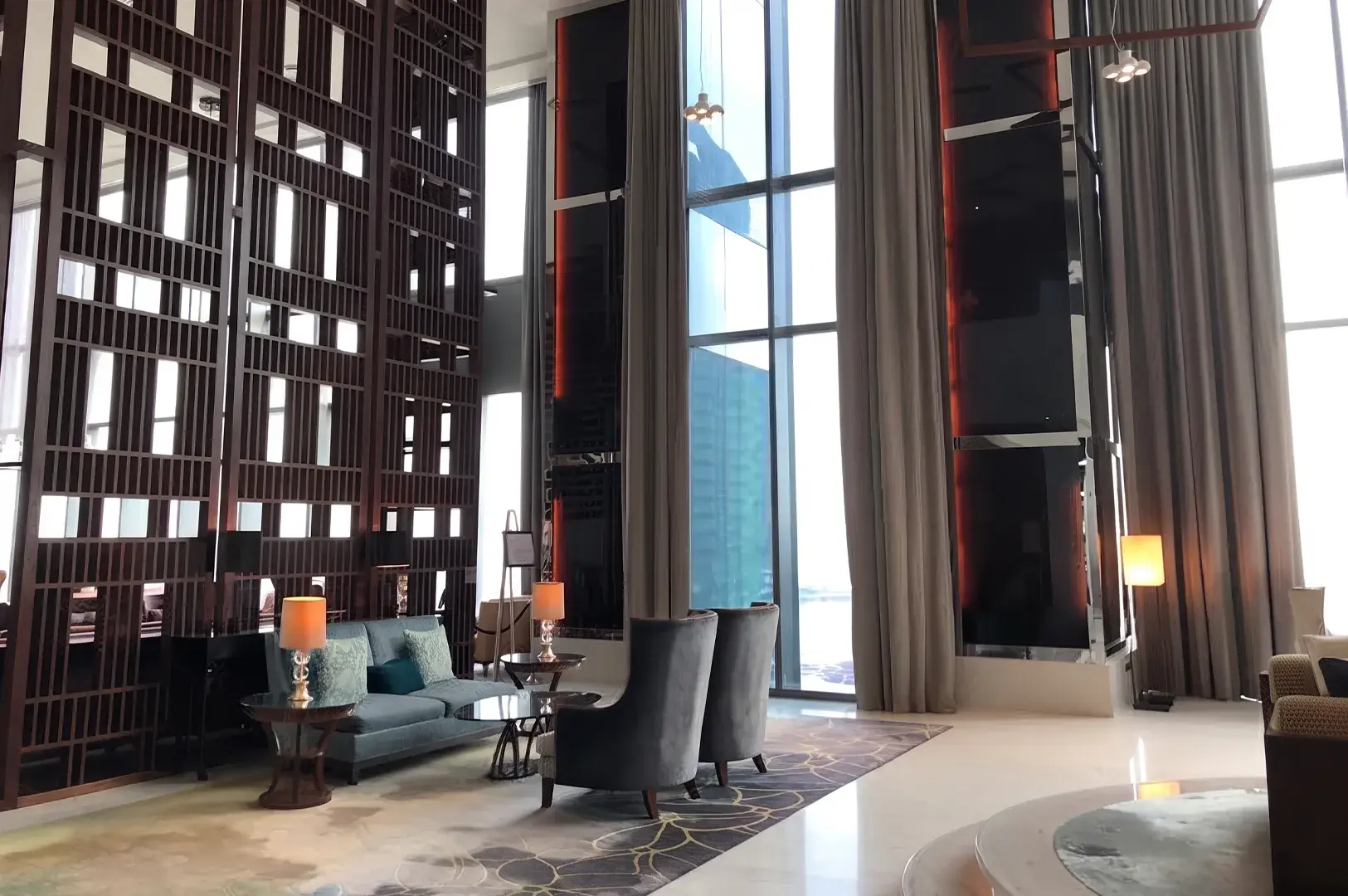 Double-height luxury lounge with tall windows, beige curtains, and dark wood lattice paneling.