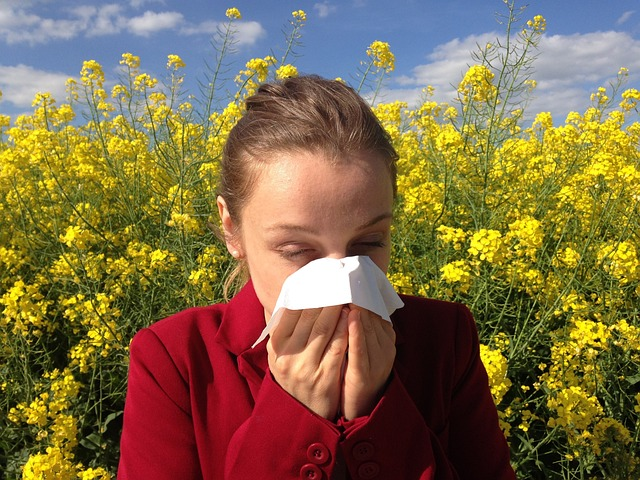 An image of a woman with seasonal allergies blowing her nose with yellow flowers behind her. 