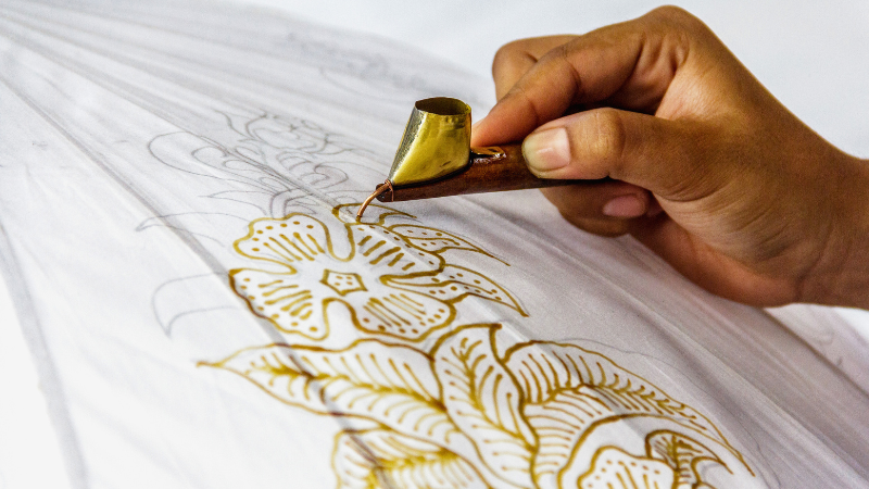 A person is using a brush to apply gold leaf to a piece of batik fabric.