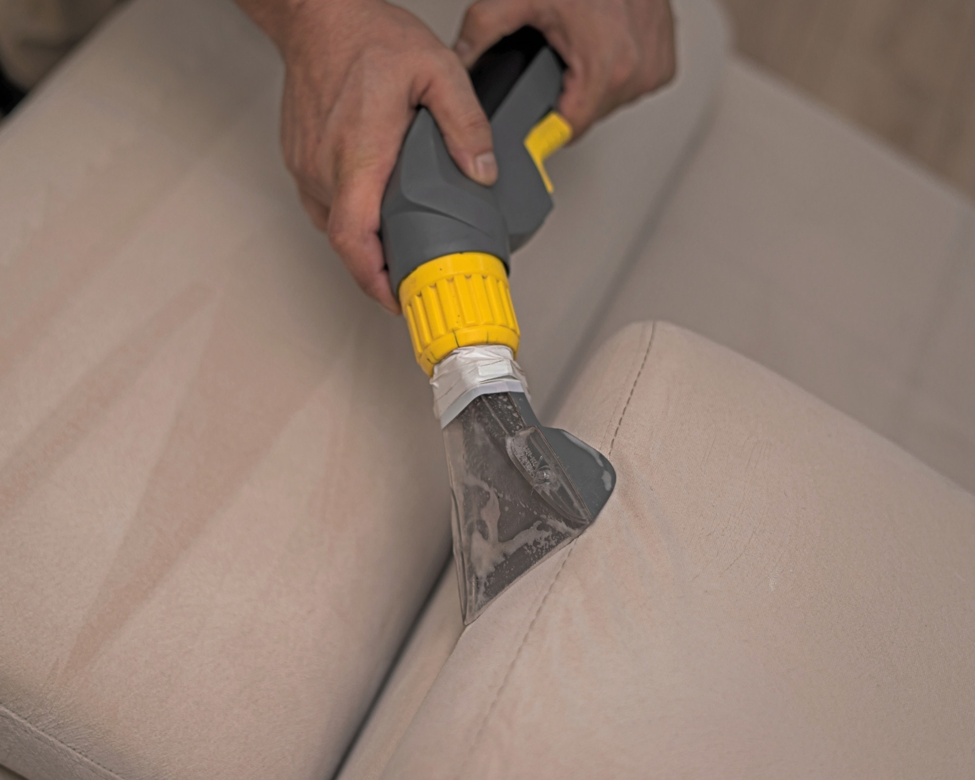 Professional upholstery cleaning on a beige sofa with a cleaning tool and brush, highlighting detailed sofa cleaning services to restore fabric furniture to its original condition.