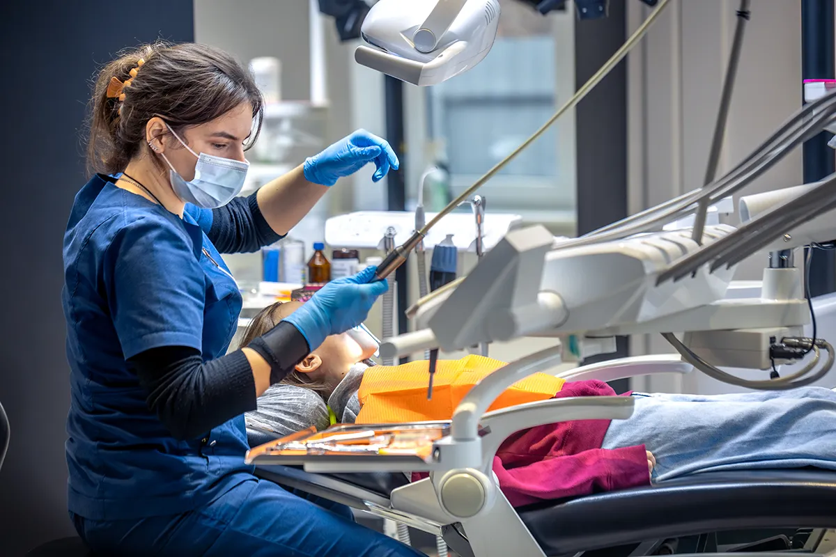 Dental hygienist performing professional teeth cleaning procedure using specialized instruments in a modern treatment room with advanced dental equipment, as part of the dental practice, overseen by a DDS.