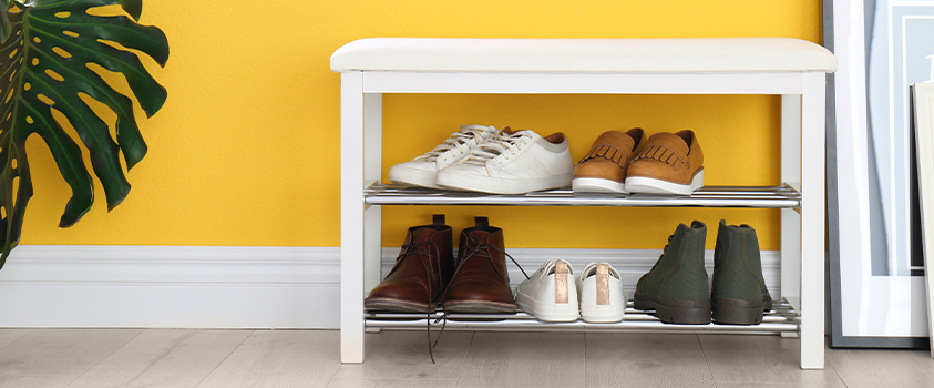 A white shoe storage bench with metal racks, holding five pairs of various shoe types. Featured against a yellow wall and light wood floor. There is a pot plant in the far left, and photo frames leaning on the far right.