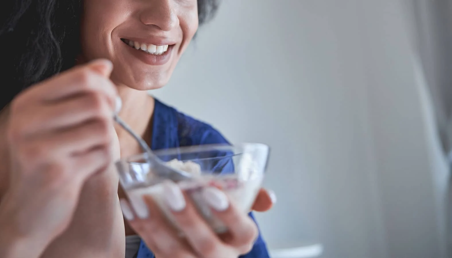 Kvinne som spiser yoghurt med en skje og smiler, sittende ved vinduet i et lyst rom.