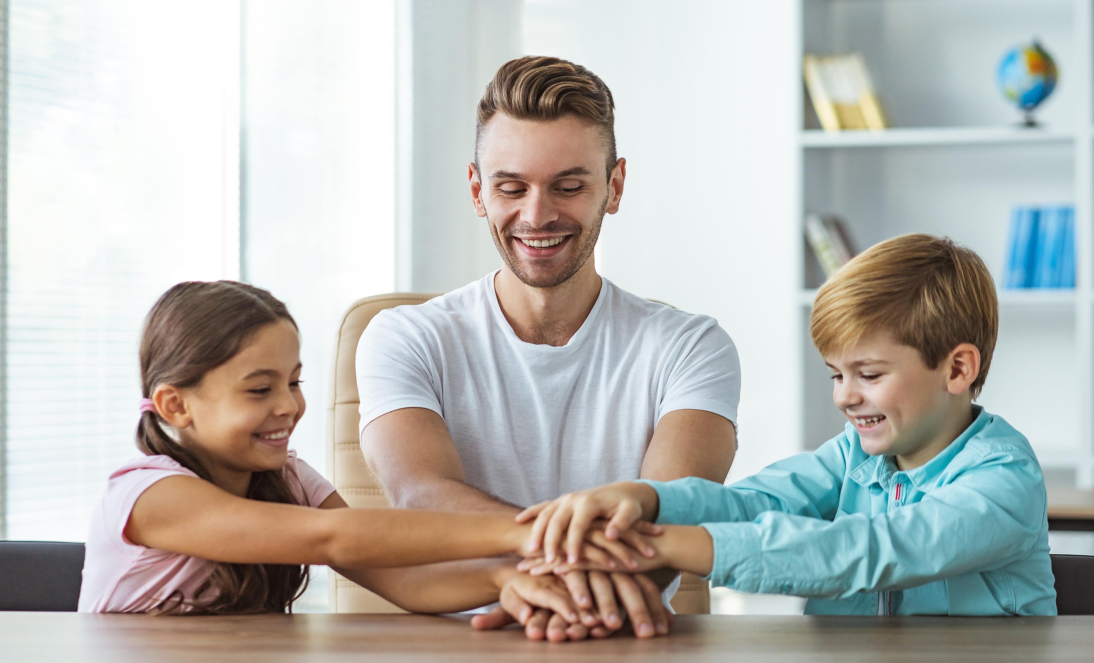 A smiling man and two cheerful children are seated at a wooden table with their hands stacked in the center.