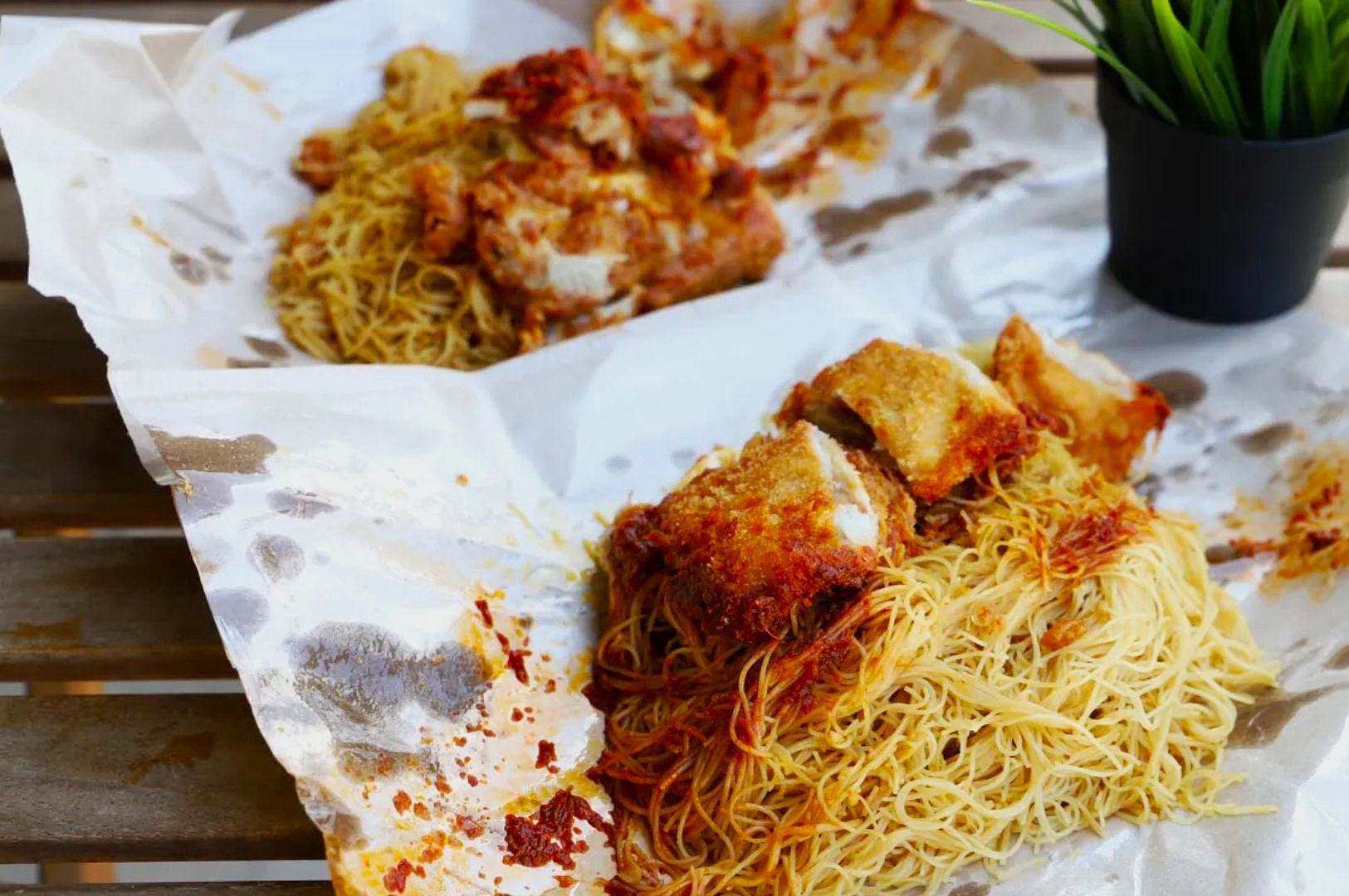 Fried noodles with crispy chicken, served on crumpled paper atop a wooden table. A small potted plant adds a touch of greenery nearby.