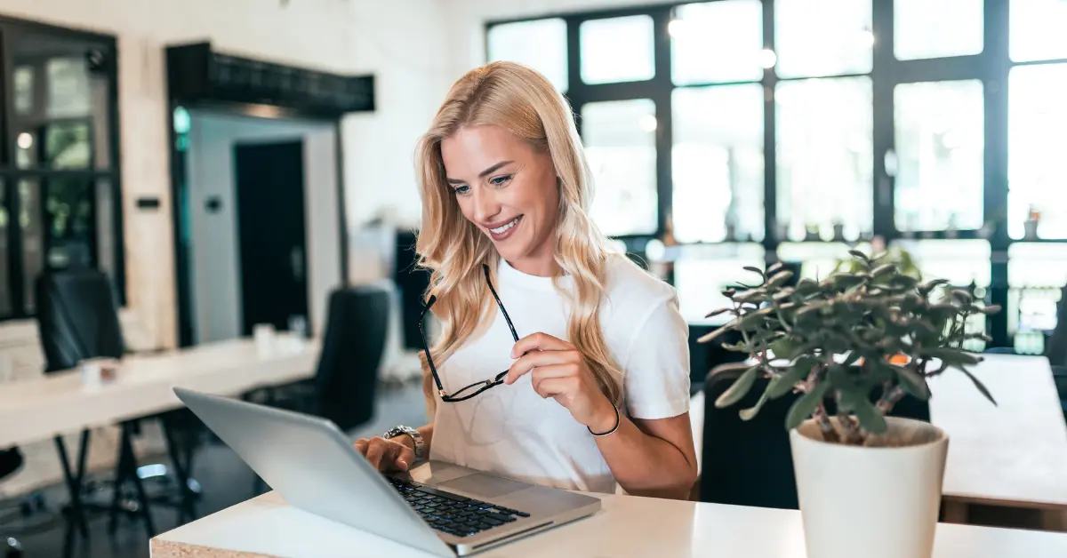 Smiling woman working on her laptop in a bright office; exploring a self-employed tax calculator.