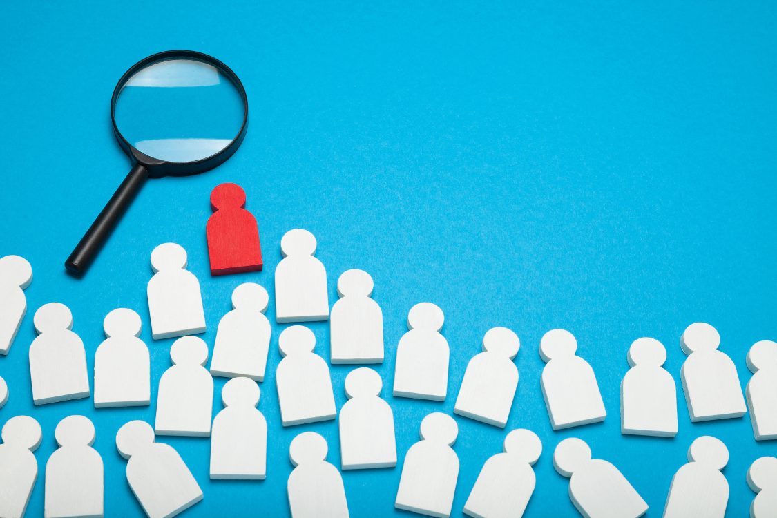 A magnifying glass highlighting a red figure among white icons symbolizing candidate selection and hiring decisions