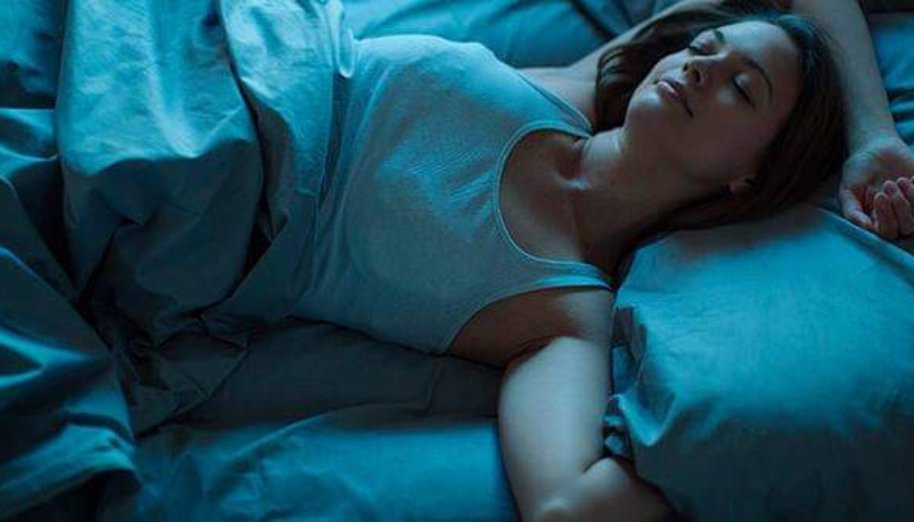 "Kvinne sover rolig i mørkt rom med blått sengetøy, armene utstrakt og ansiktet avslappet i behagelig sovestilling."