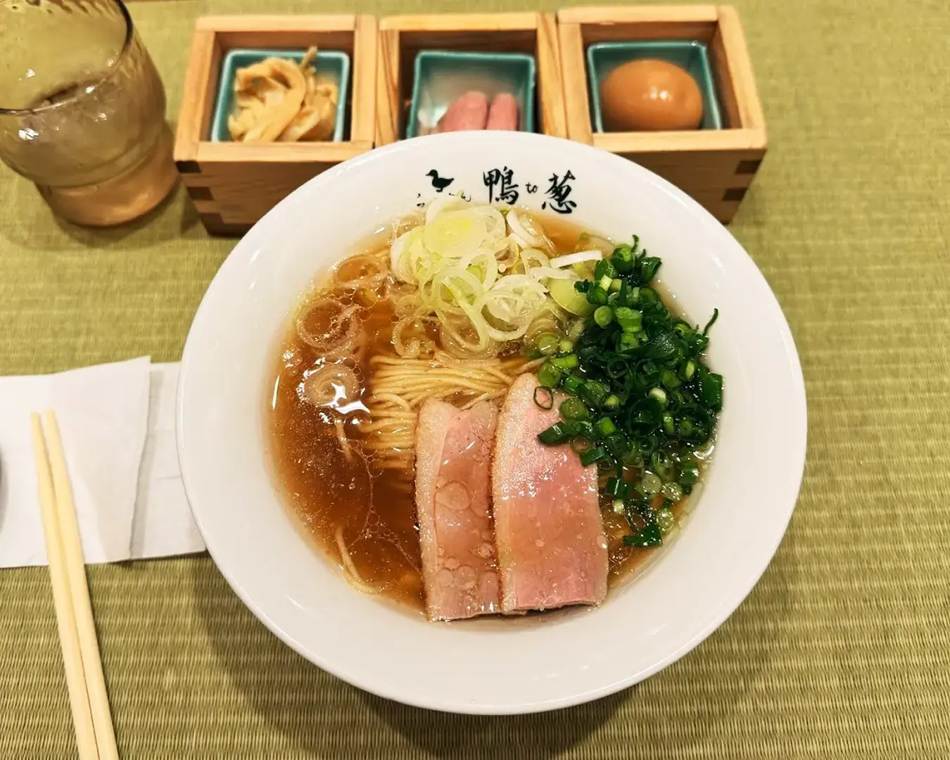 A bowl of ramen with sliced pork, chopped green onions, and leeks in broth, on a green mat. Side dishes include an egg and pickled items.