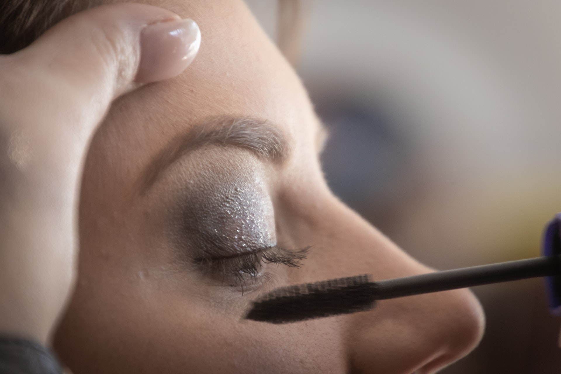 Detalhe mostrando a mão de um maquiador prestes a aplicar uma máscara de cílios no olho de uma jovem mulher. Não é possível ver muitos detalhes do seu rosto, mas sua pálpebra está coberta com uma sombra prateada brilhante, e sua pele é clara. Imagem: Magda Ehlers, Pexels.com.