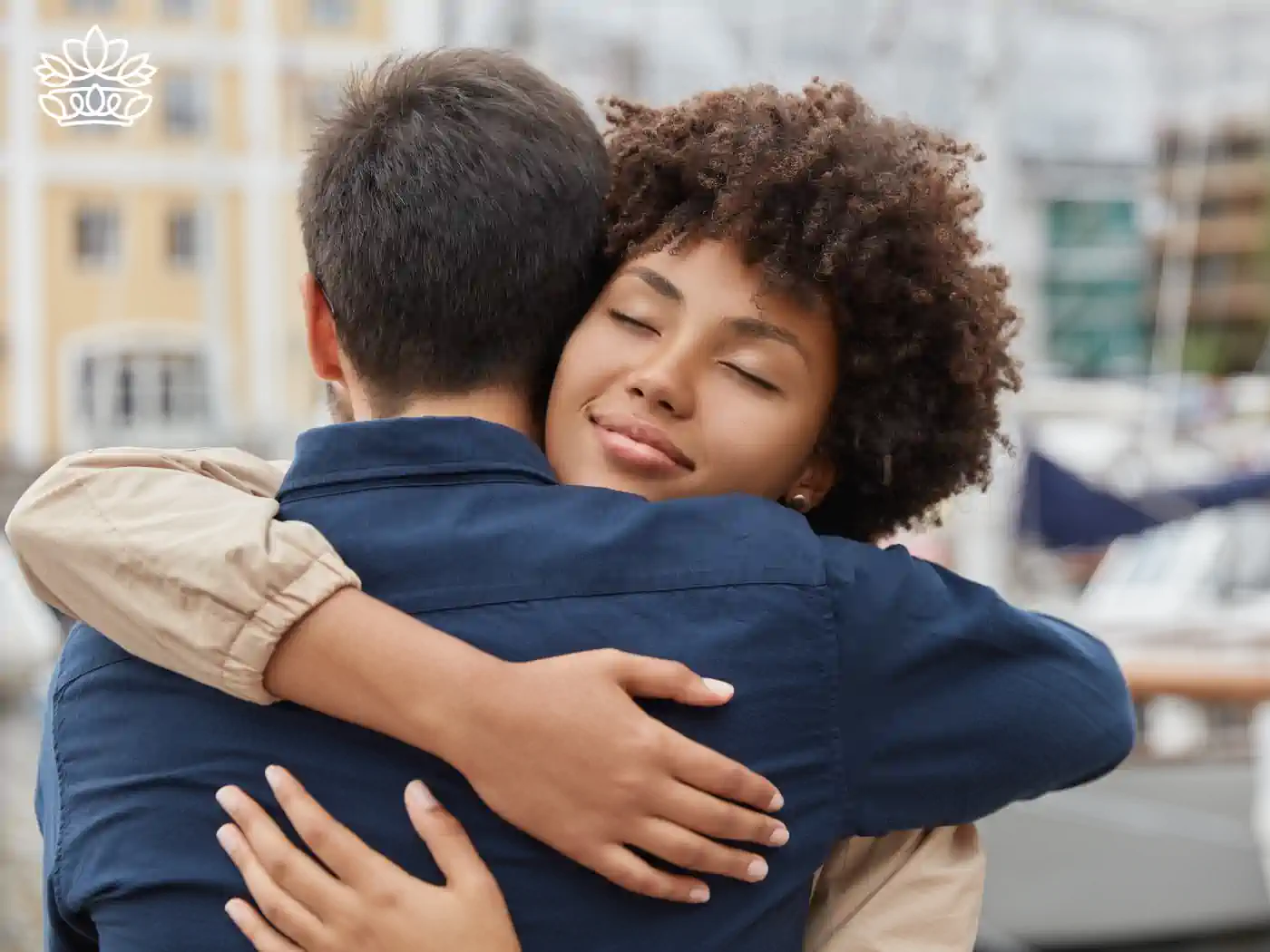 Warm embrace between a young woman with curly hair and a man, sharing a comforting moment in a serene marina setting. Coffin sprays from Fabulous Flowers and Gifts.