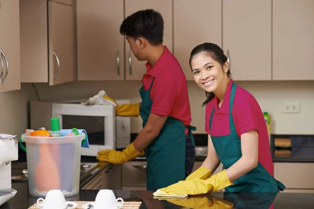 Two professional cleaners wearing matching uniforms and yellow gloves cleaning a modern kitchen — one person wipes a microwave while the other smiles at the camera while polishing a countertop