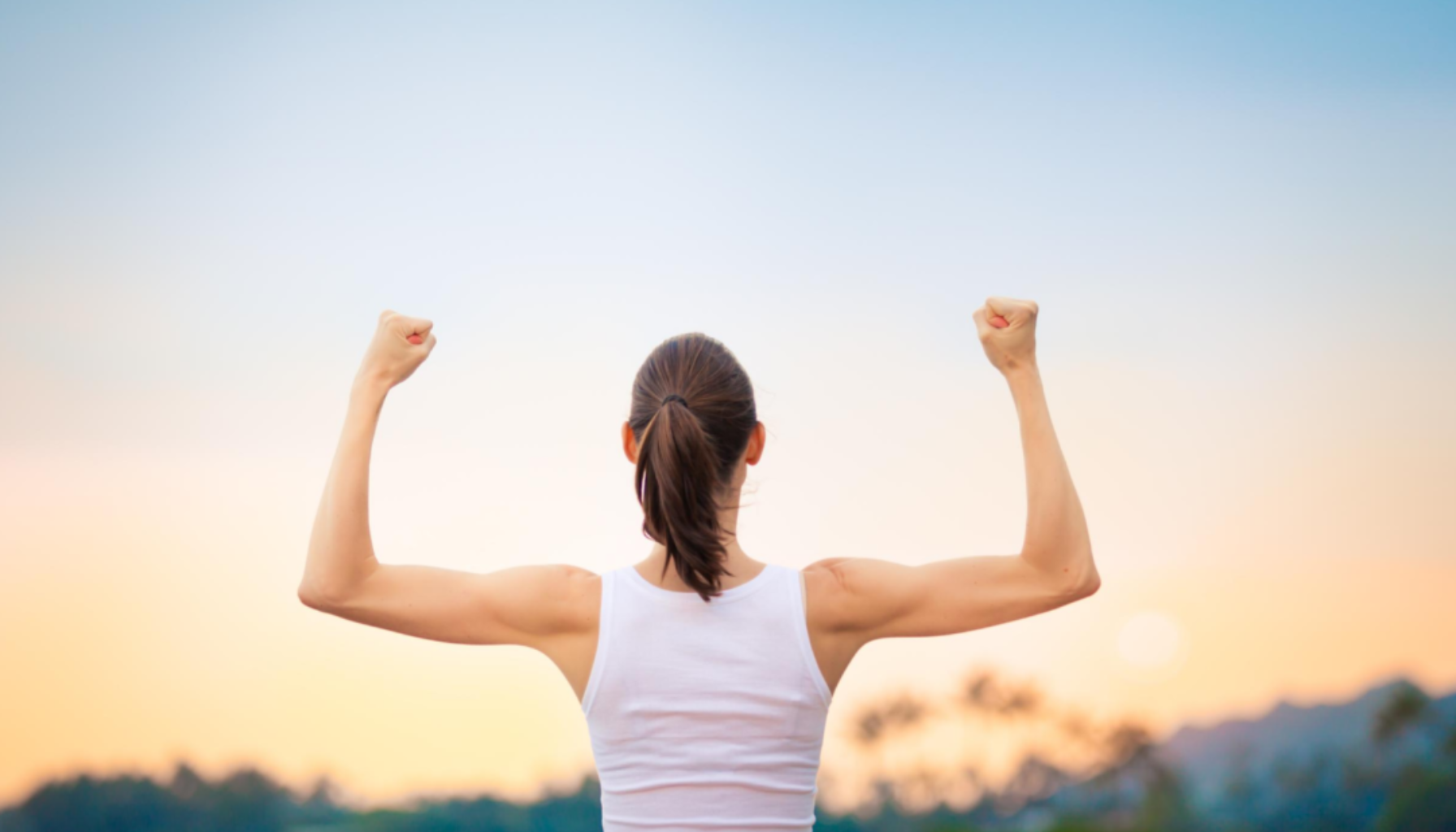 Person sett bakfra løfter armene i seiersposisjon utendørs mot lys himmel og rolig landskap.