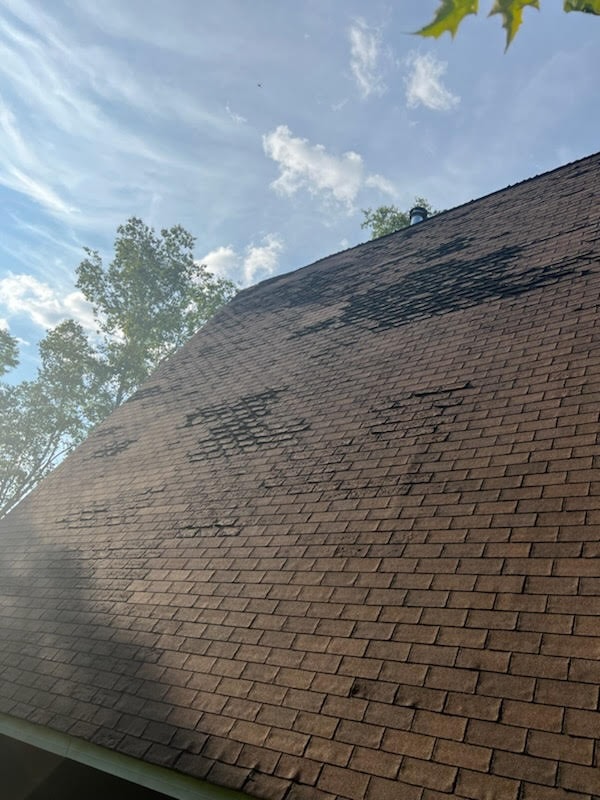 A picture of curling and peeling shingles in the summer with green trees and a blue sky with clouds in the background.