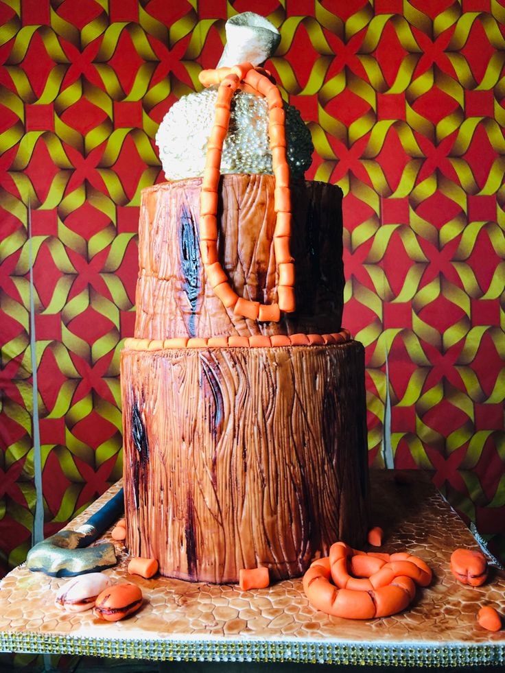 A two layered cake with a wooden look and feel surrounded by red beads, cowries, and an axe