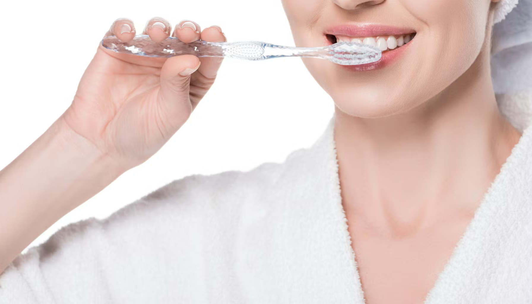 Helse og munn: Hvordan kroppens tilstand påvirker tennene 15 Kvinne i hvit morgenkåpe pusser tennene med gjennomsiktig tannbørste, smiler mens hun utfører daglig munnhygiene.