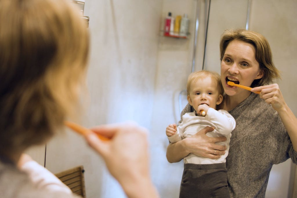 Uma mulher está escovando os dentes em frente a um espelho no banheiro enquanto segura um bebê no colo. O bebê, que tem cabelos claros e usa um body branco com detalhes em marrom, observa a cena atentamente, com uma mãozinha próxima à boca, como se imitasse o gesto de escovar os dentes. A mulher, com o cabelo preso e uma expressão concentrada, olha para o espelho enquanto escova os dentes com uma escova de dente laranja. O ambiente transmite um momento cotidiano de cuidado pessoal, onde a mulher consegue conciliar a sua higiene bucal com o cuidado do bebê.