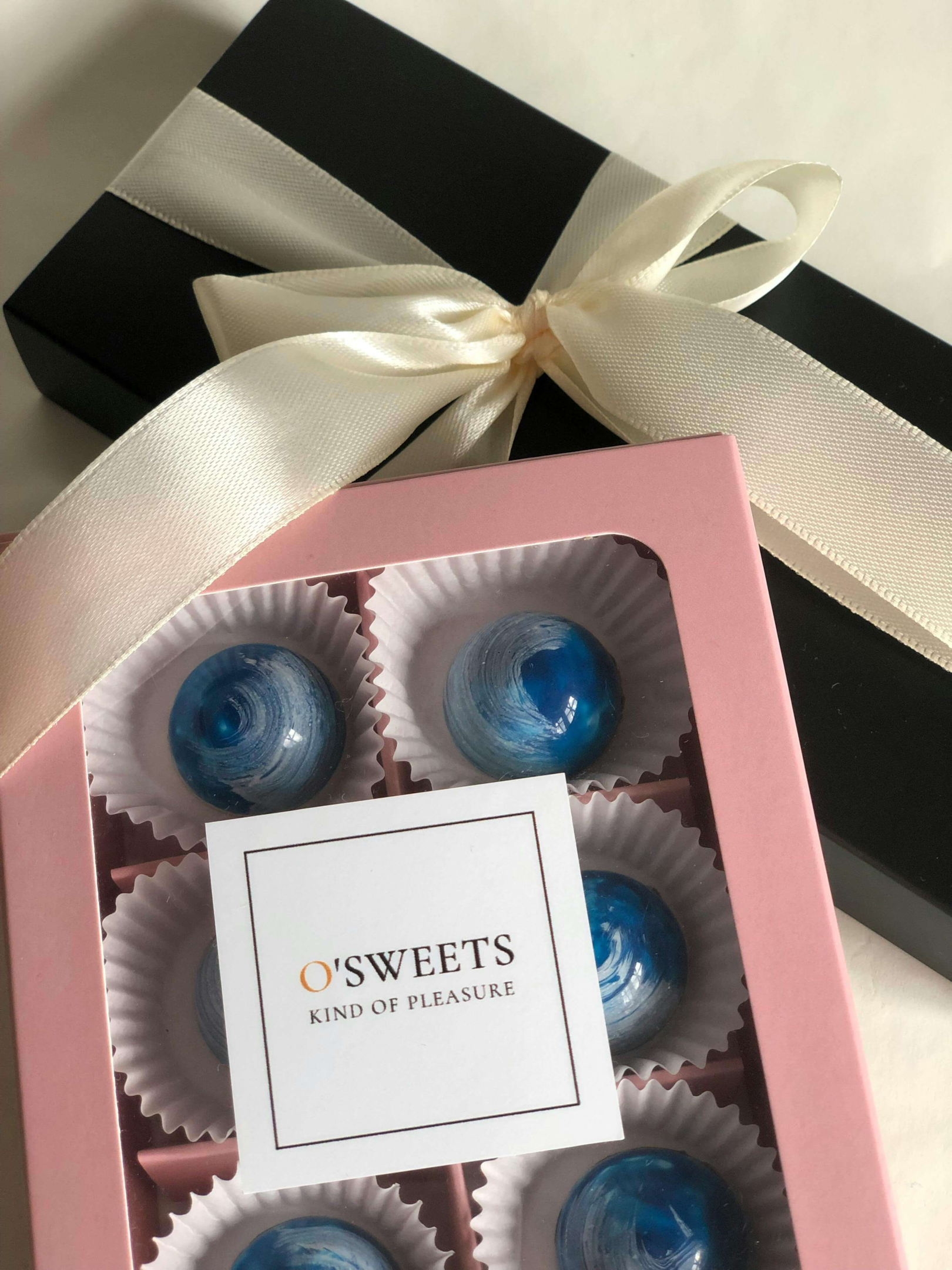 Pink box of glossy blue marbled chocolates with a white ribbon-tied black gift box on a light background, labeled 'O'Sweets Kind of Pleasure'