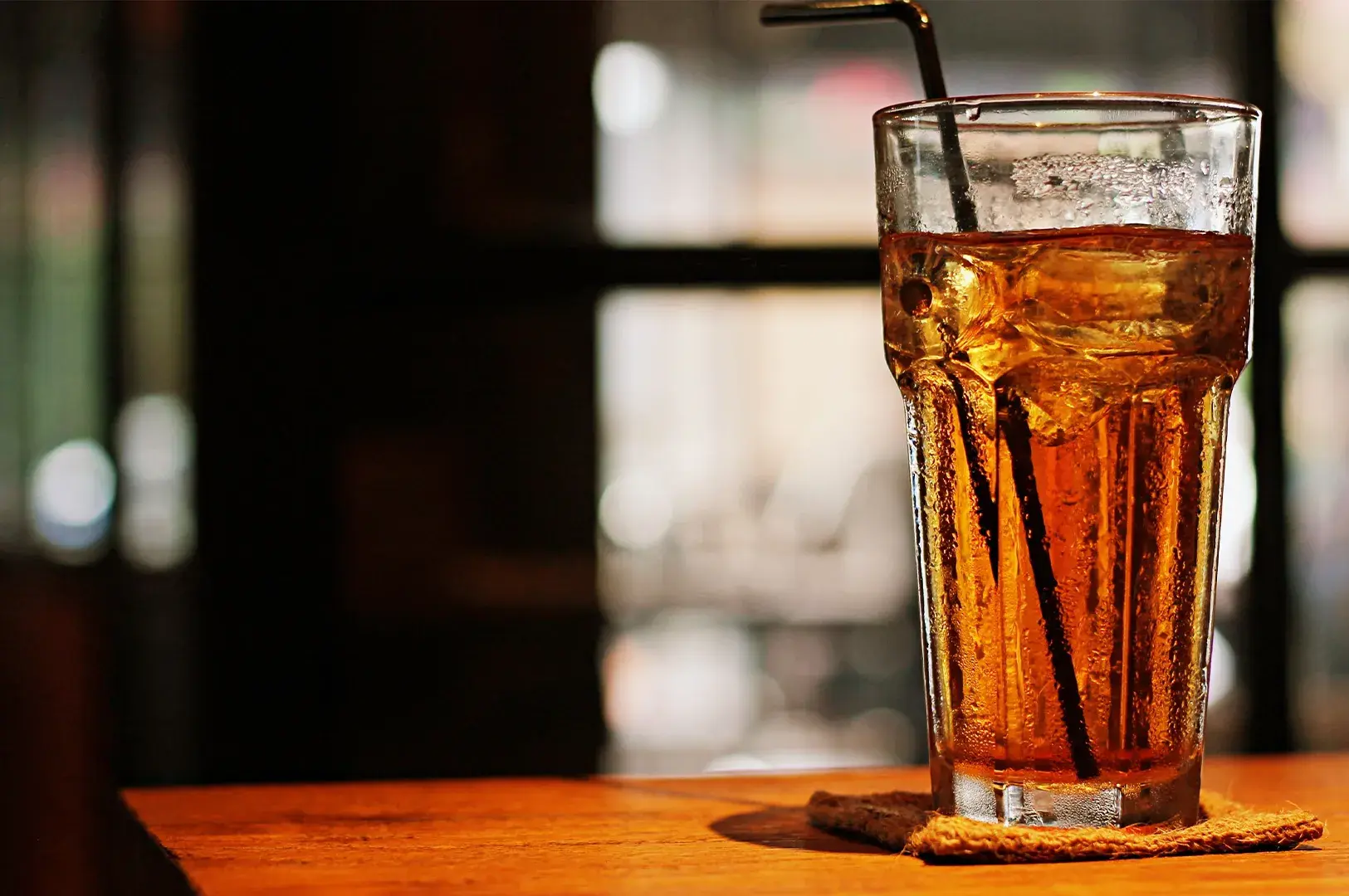 Tall glass of iced black tea with ice and a straw on a coaster.