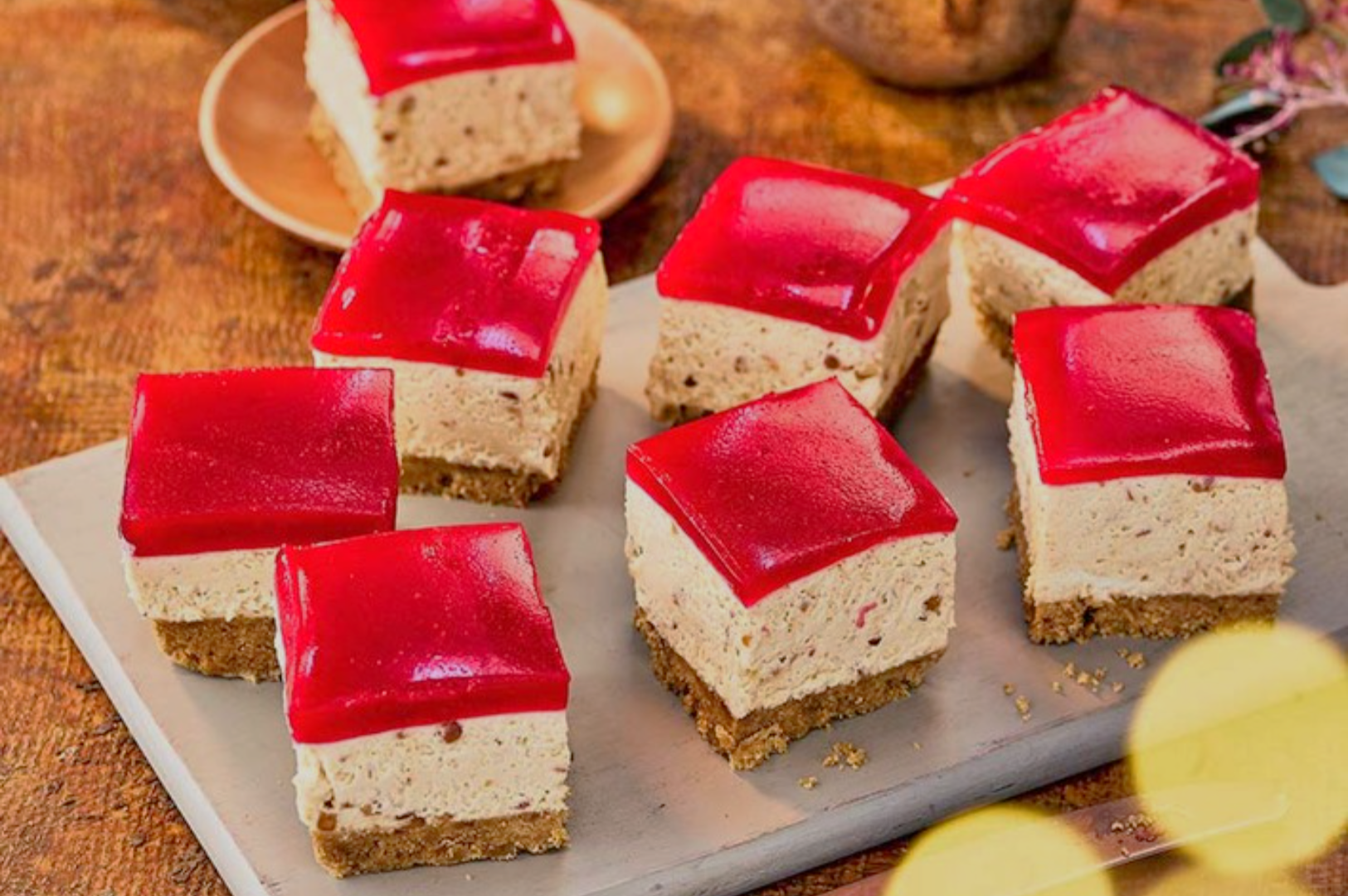 Eight cheesecake squares with red jelly tops are arranged on a beige platter. The creamy and crumbly textures are highlighted against a rustic background.