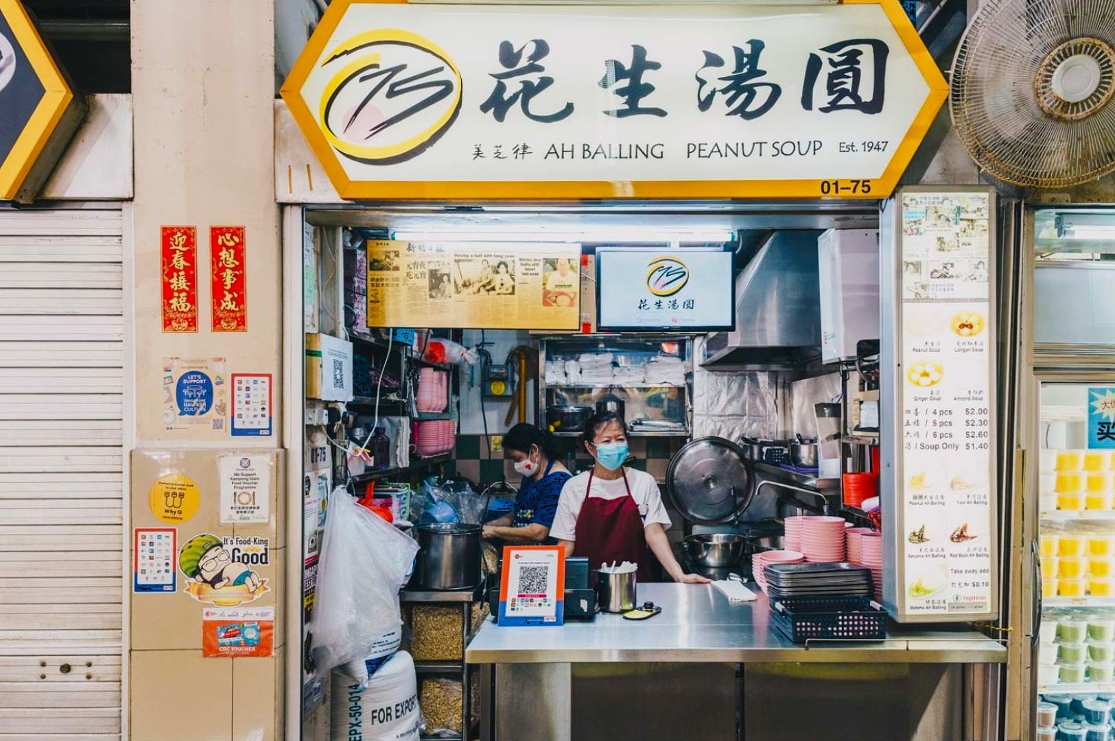 A bustling hawker stall named "Ah Balling Peanut Soup." Two staff members wearing masks work inside, surrounded by cooking utensils and ingredient containers.