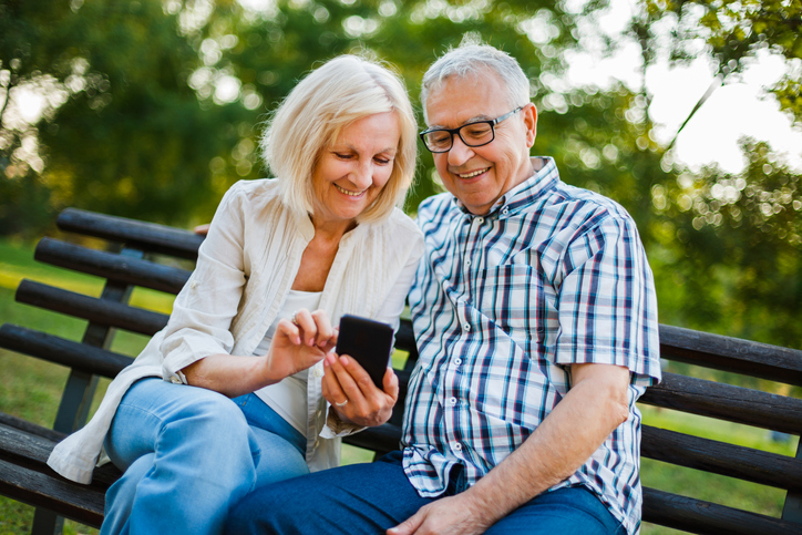 Happy older couple sitting on a park bench with a cell phone. 