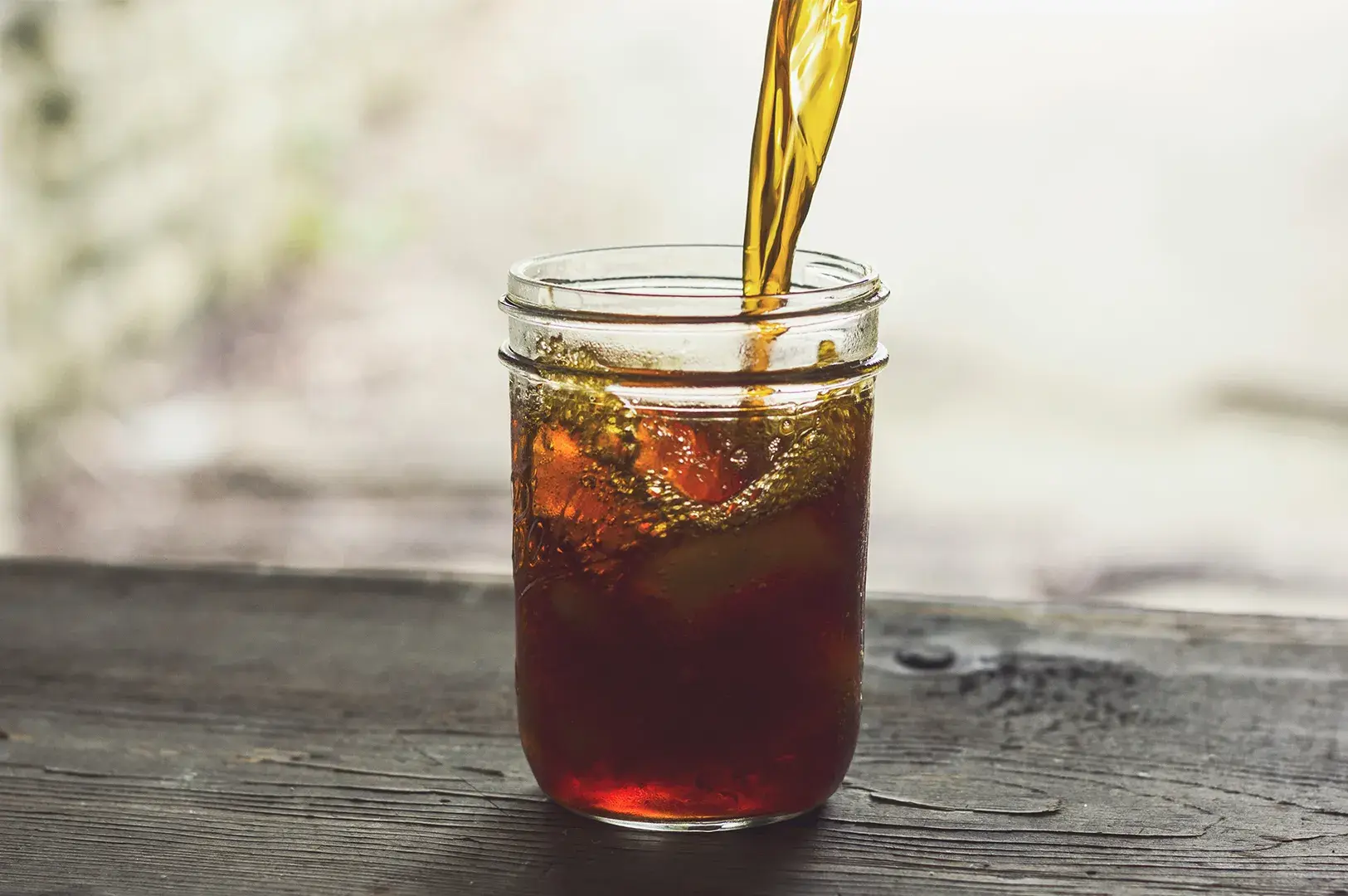 Iced tea beverage pouring into a mason jar with ice on a wooden table.