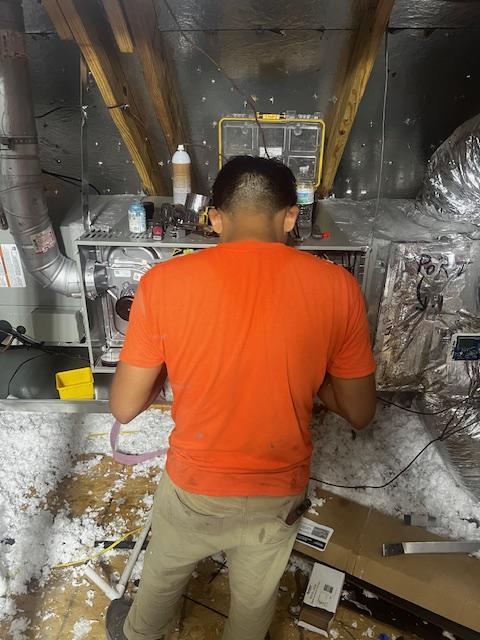 An HVAC technician wearing a navy shirt is installing a Trane air conditioning system in a residential attic space, focusing on the indoor unit while ensuring proper ductwork placement. This installation aims to enhance energy efficiency and improve the home's cooling needs, ultimately leading to long-term savings on energy bills.