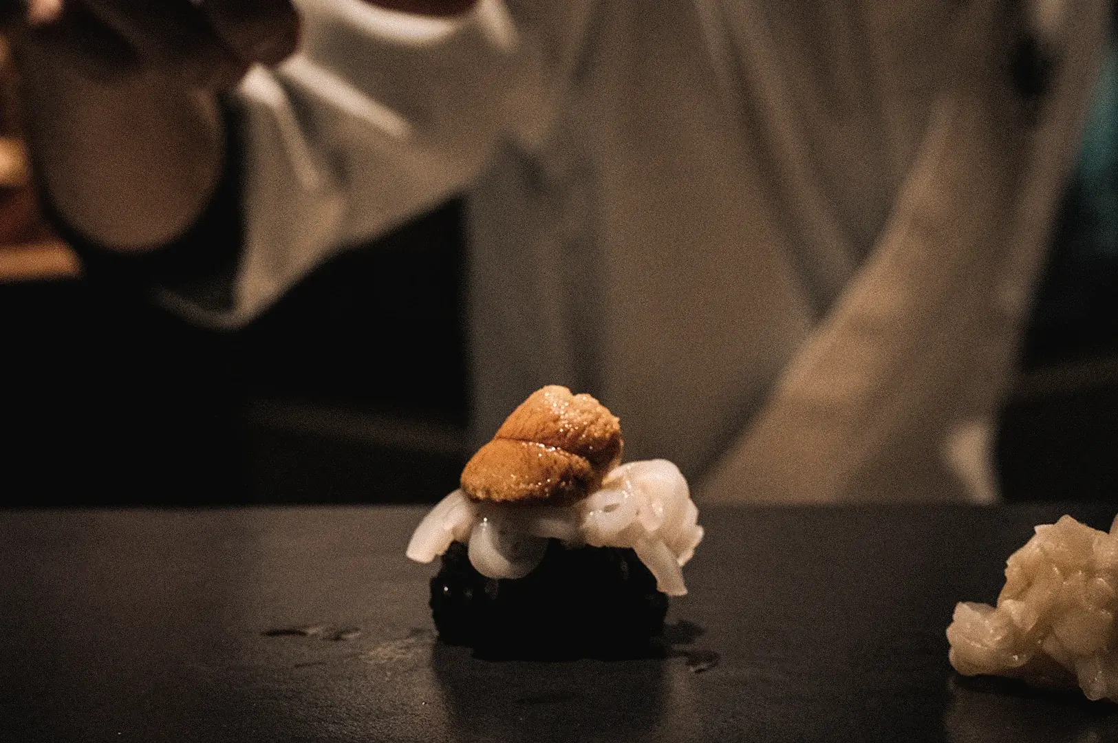 A close-up macro shot highlights a freshly prepared piece of squid and sea urchin nigiri resting on the counter at Sushi to Sumi standing sushi bar.