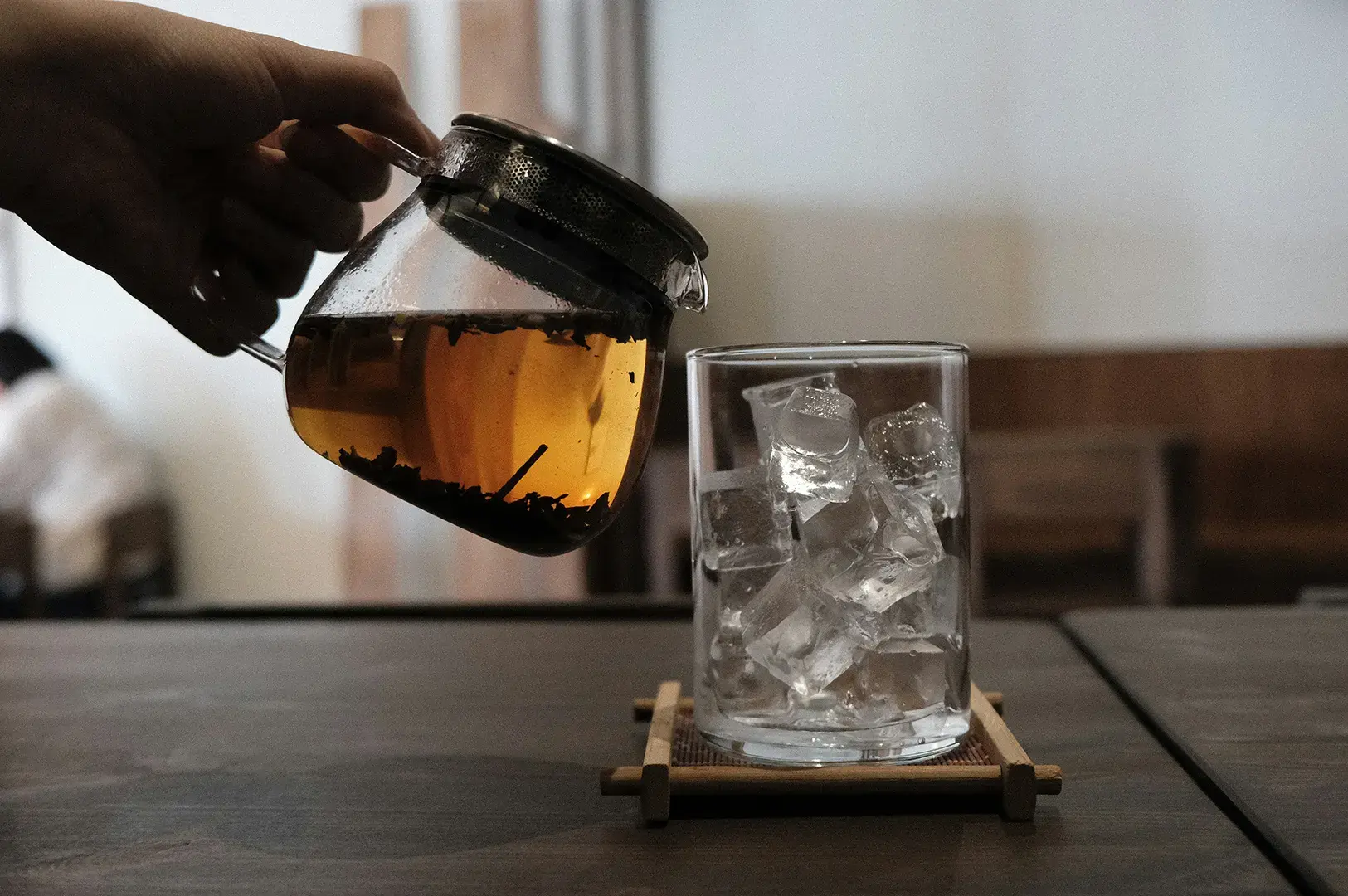 Person pouring brewed tea from a glass teapot with a built-in strainer into an empty glass filled with ice cubes on a wooden table.