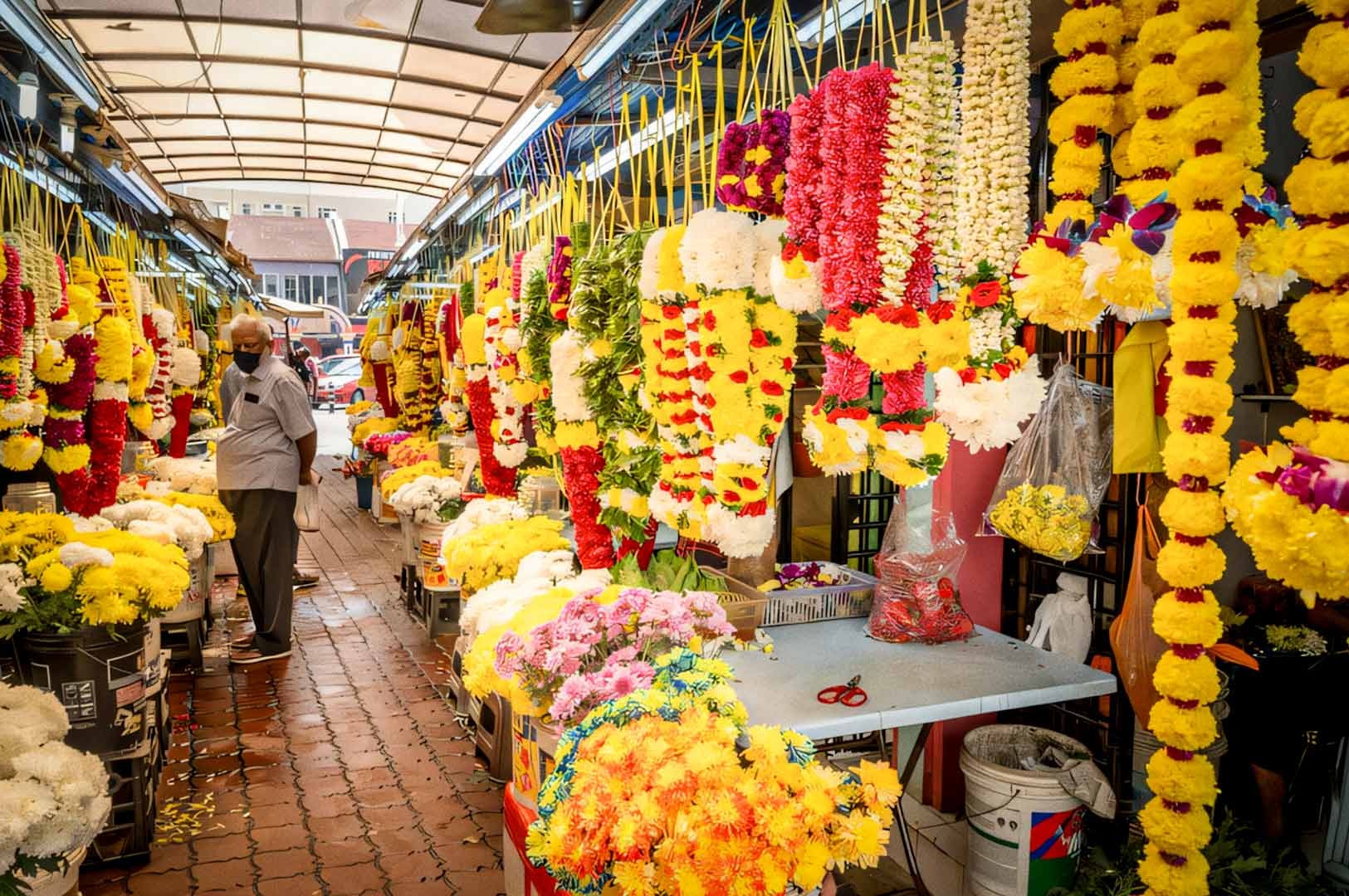 A vibrant aisle in Tekka Market filled with colorful flower garlands and fresh blossoms hanging and displayed along both sides.