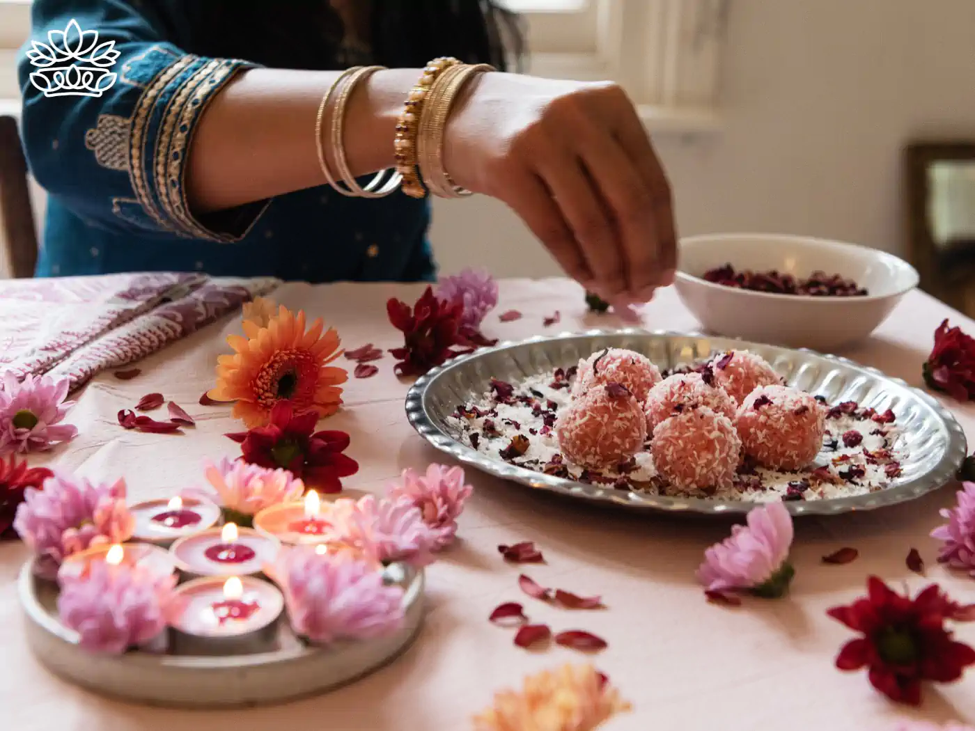 A woman in traditional attire preparing for Diwali by arranging a tray of coconut sweets and lighted tealight candles surrounded by fresh flowers on a decorated table. Fabulous Flowers and Gifts - Diwali Flowers. Delivered with Heart.