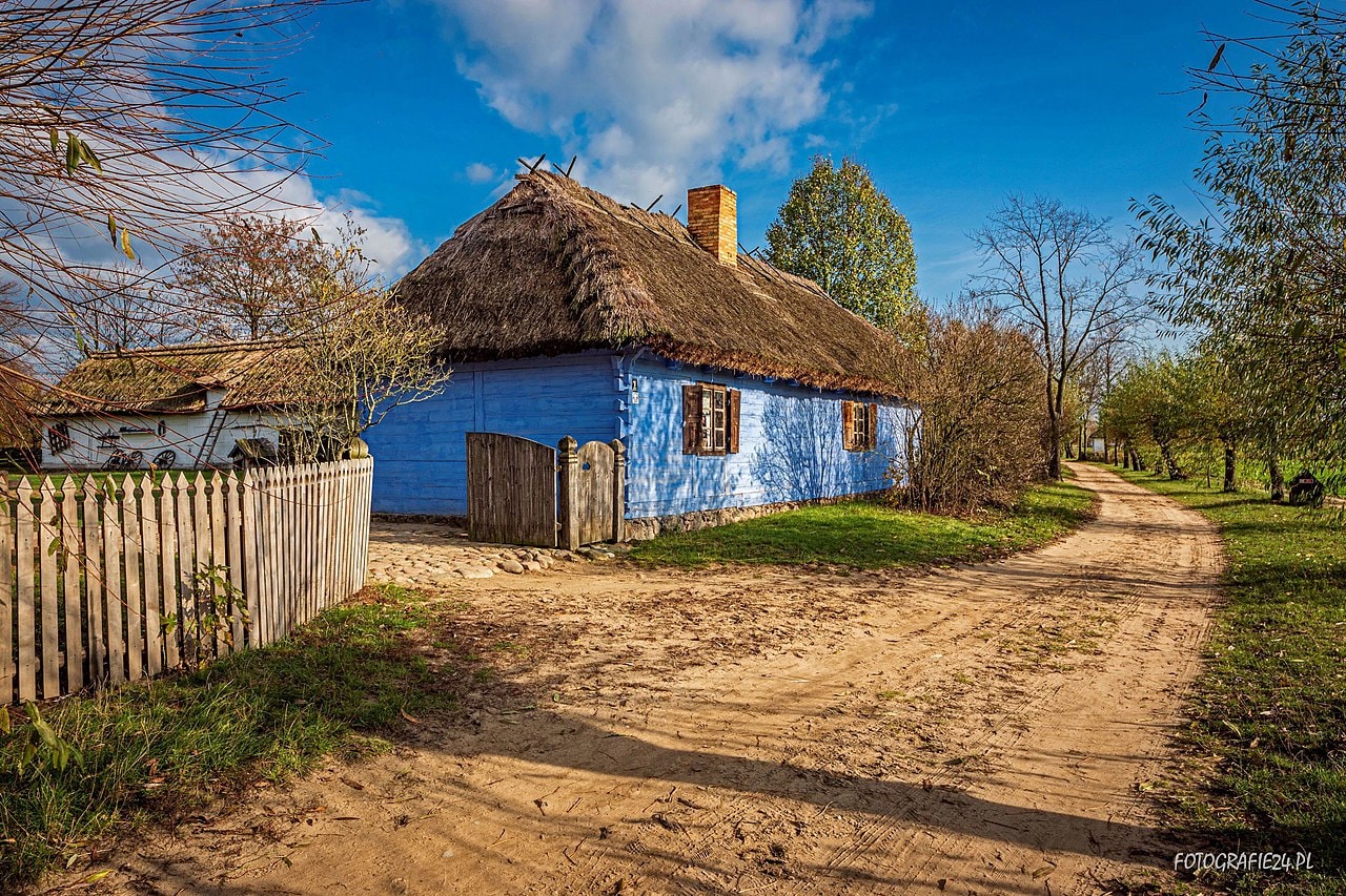 Tradycyjna, drewniana chata kryta strzechą, otoczona drewnianym płotem, z ogrodem pełnym zieleni. (źródło:https://commons.wikimedia.org/wiki/File:Zagroda_z_Izdebna_Ko%C5%9Bcielnego_-_Muzeum_wsi_mazowieckiej_w_Sierpcu.jpg)