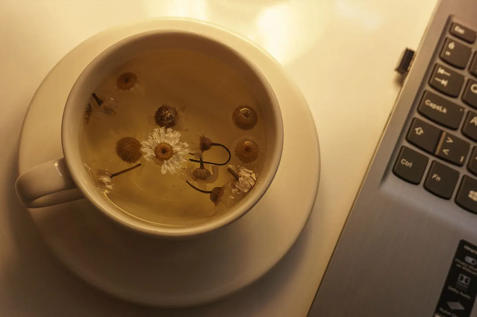 An overhead, top-down view shows a simple white ceramic cup and saucer filled with translucent golden chamomile tea, where several whole chamomile flowers drift on the surface. The tea setting sits on a bright white desk directly adjacent to the right side of a sleek, silver laptop keyboard, with keys like Control, Function, and Caps Lock visible. The scene is bathed in a warm, amber-toned glow, evoking a calm and comforting mood during a work break.