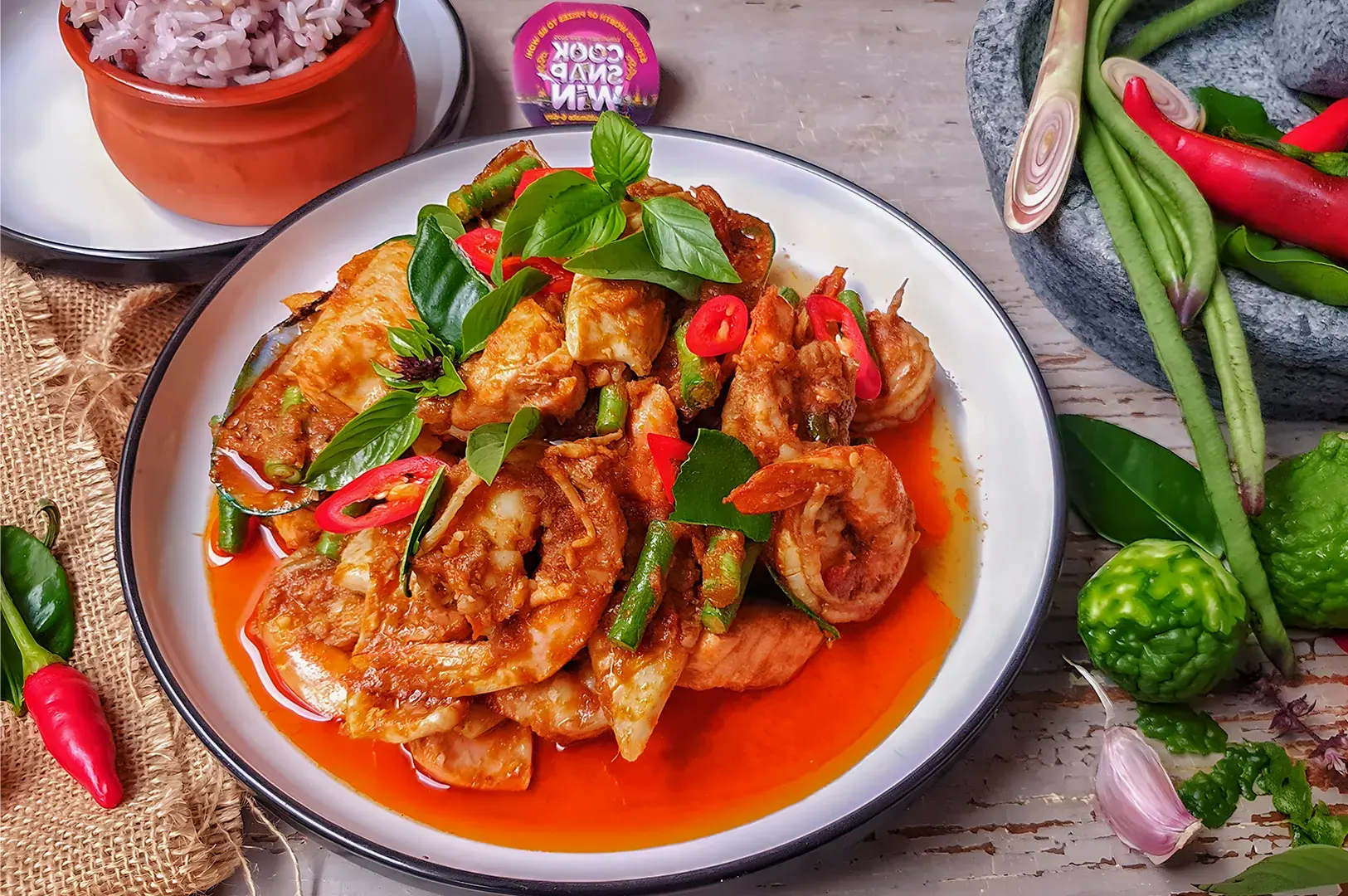 Spicy shrimp dish garnished with basil and chili slices on a white plate. Served with purple rice in a rustic bowl. Fresh ingredients nearby exude a vibrant, appetizing look.