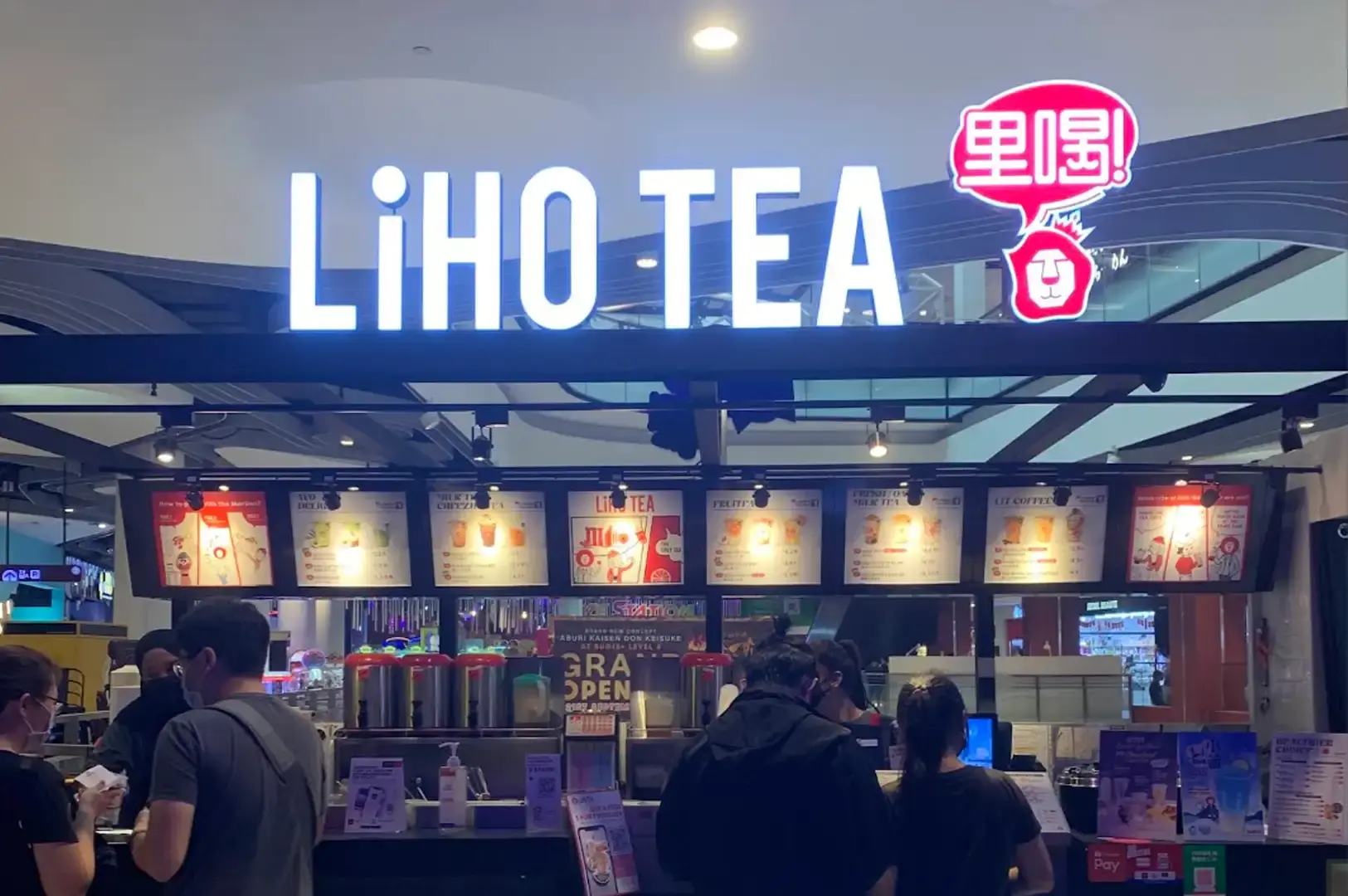A brightly illuminated LiHO Tea store counter inside a shopping mall, featuring a large glowing white brand sign above a red lion speech bubble logo, digital menu boards highlighting seasonal tea offerings, and patrons ordering at the service counter.