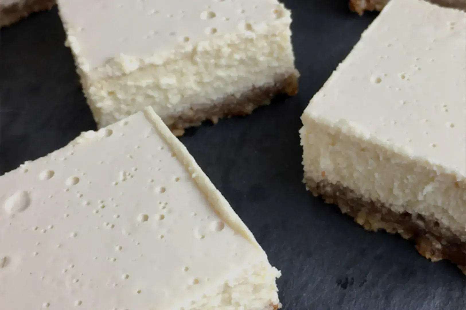 A close-up of square pieces of cheesecake with a graham cracker crust on a dark surface.